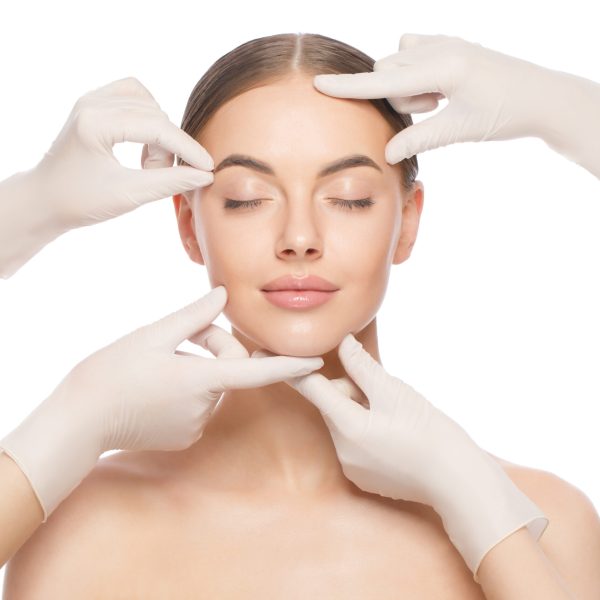 close up portrait of young woman standing with naked shoulders and neck, face is touched by beauticians in gloves, preparing her for plastic surgery procedures, isolated on white background